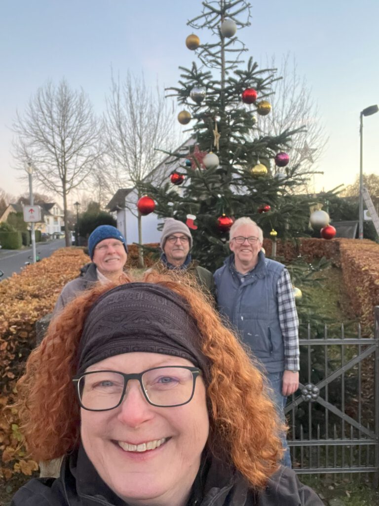 Vier Menschen, eine Frau im Vordergrund, drei Männer im Hintergrund vor einem geschmückten Weihnachtsbaum.